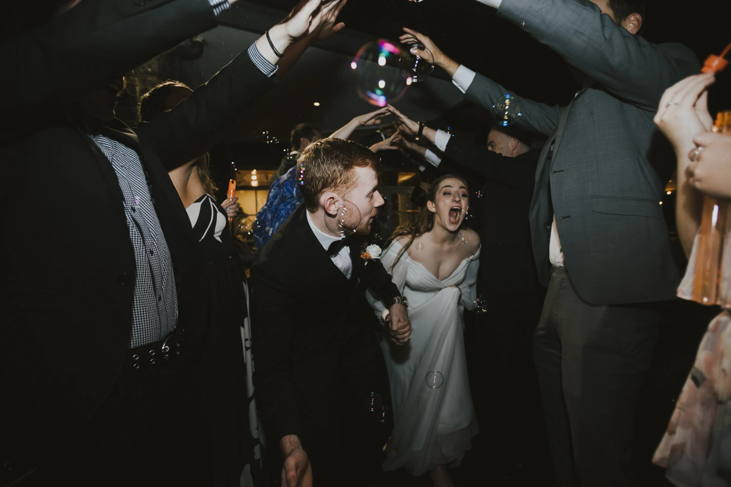 A newlywed couple running through a tunnel of raised arms and bubbles