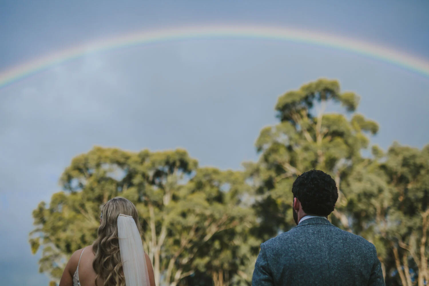 Newlyweds seen from behind beneath a full rainbow over country trees